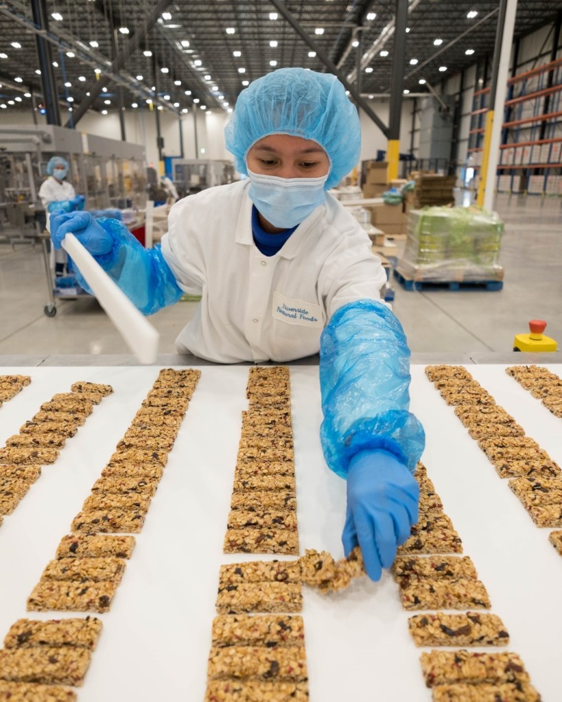 A person in a white coat and blue gloves that prepares the granola made of the Madegood grass on a large table.