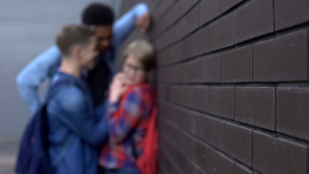 Group of students harassing a classmate in a backyard, the person bending over a brick wall with 'dumb donut' written on the forehead